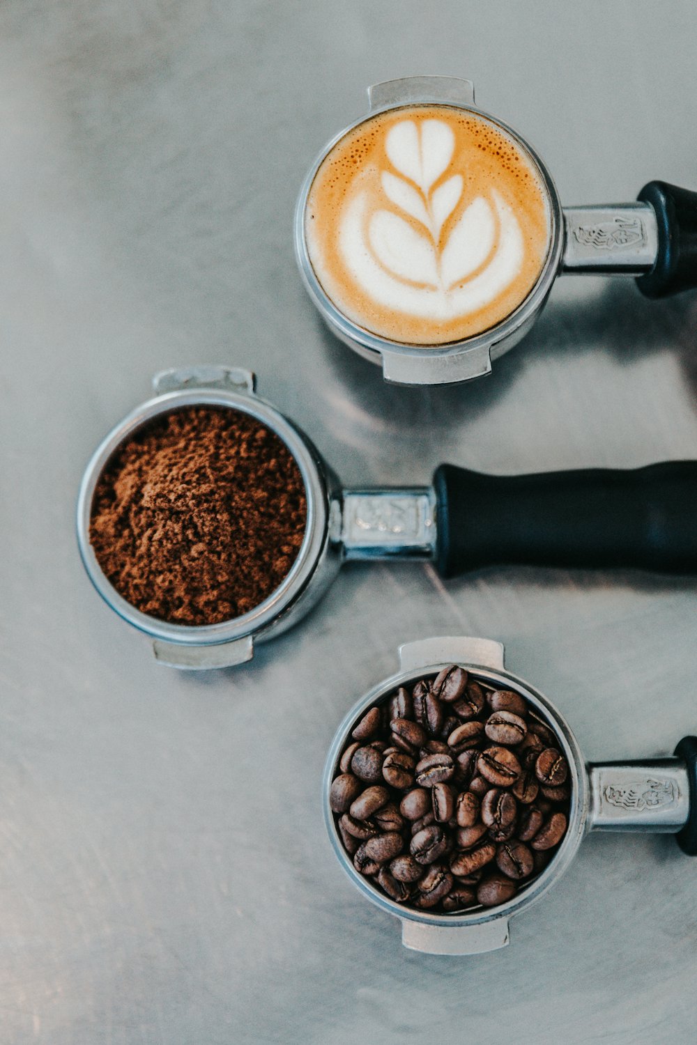 Three portafilters on a concrete counter: whole beans, ground coffee, and a finished cup.